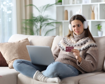 woman sitting on couch wearing headphones and making strange face at credit card and laptop