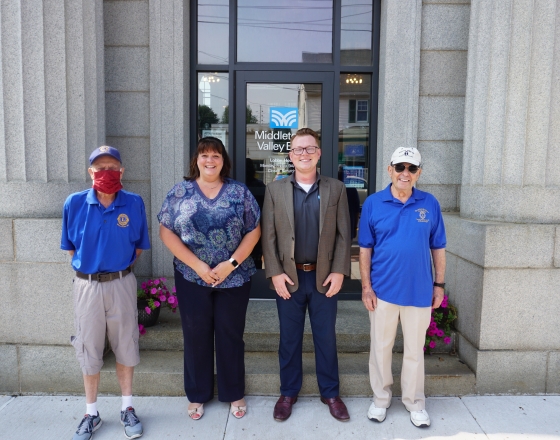 two men in blue shirts on left and right and woman and man in middle in front of building