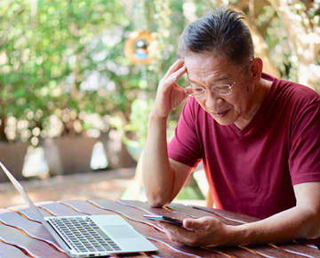 man in red shirt holding head and looking down at phone and laptop